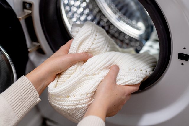Woman putting white sweater clothes into the drum of a washing machine, front view.