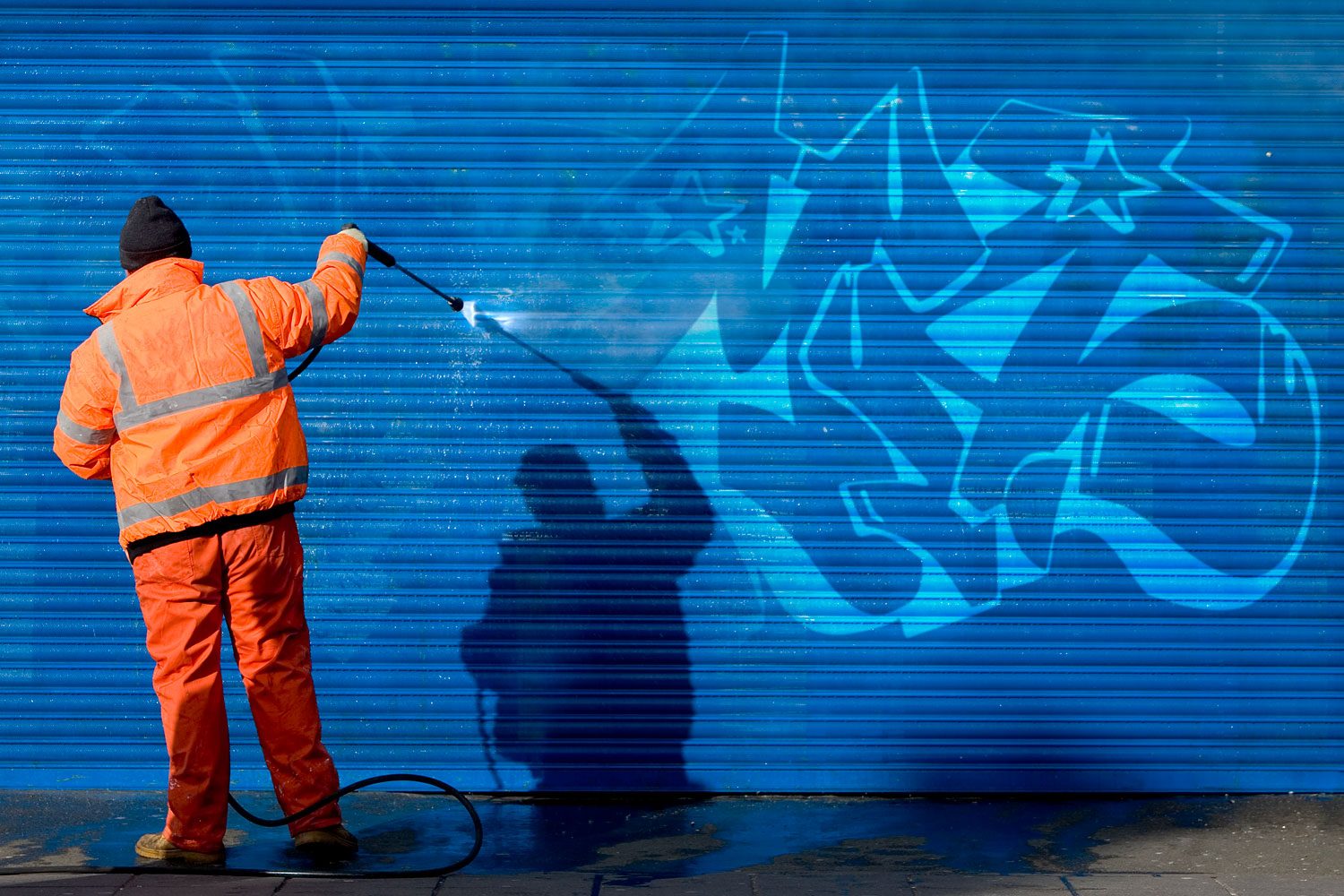 Worker in Reflective Clothing Washing Graffiti Off A Blue Security Grill