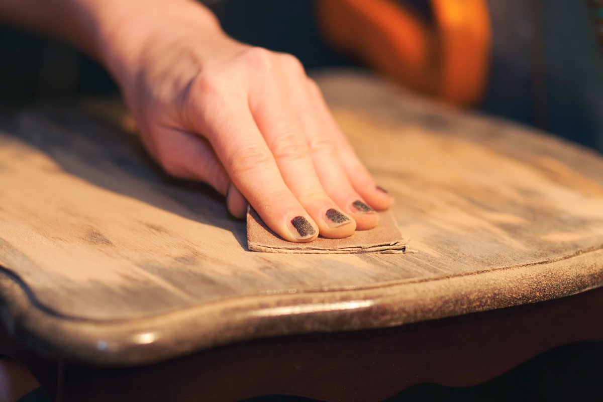 Woman's Hand Works to Sand Finish off of a Wooden End Table By Using Sandpaper, covering her hands with the removal of the varnish