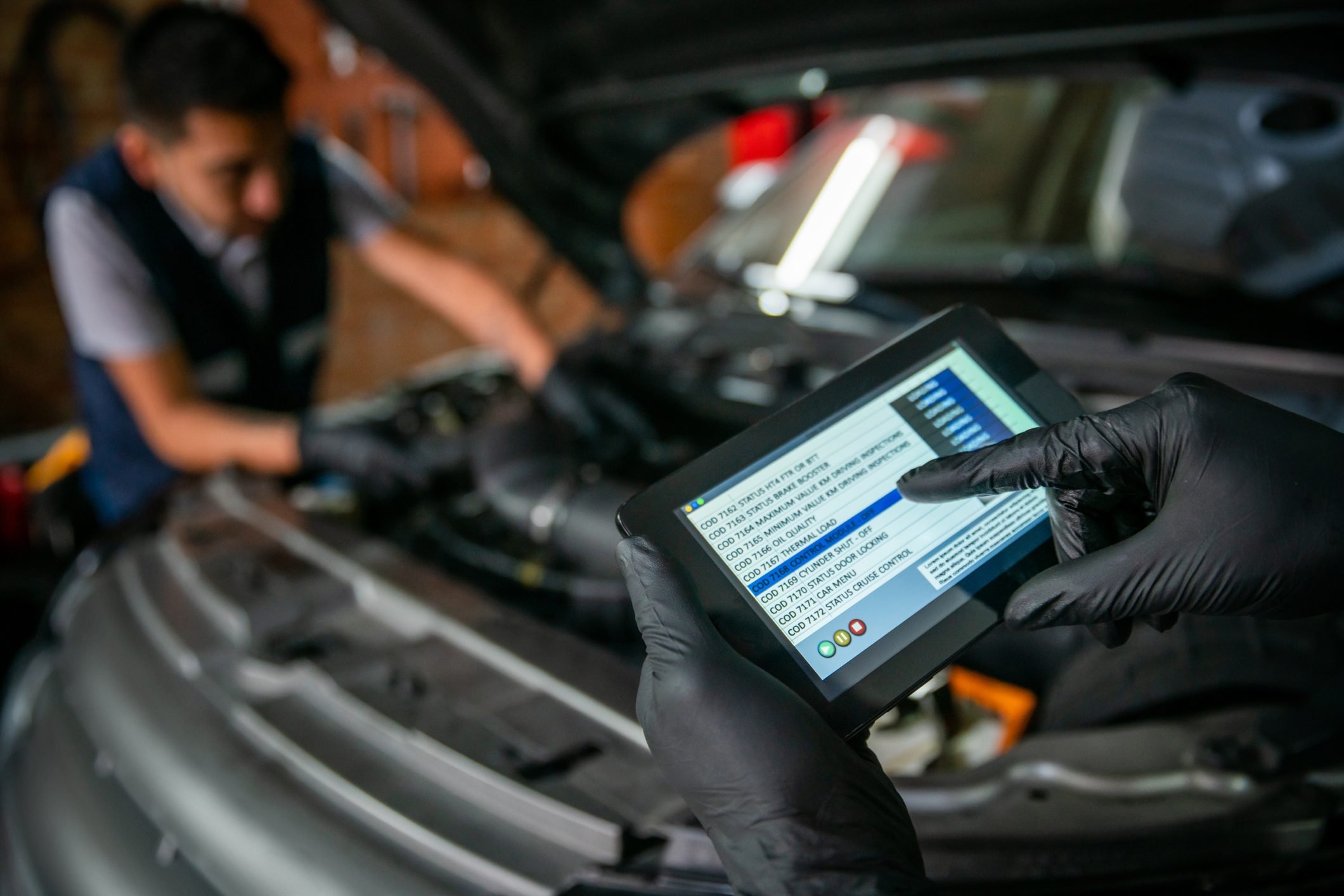 Team of mechanics working together on a car, one of them holding a tablet to check the sensor readings while the other one adjusts them on the engine