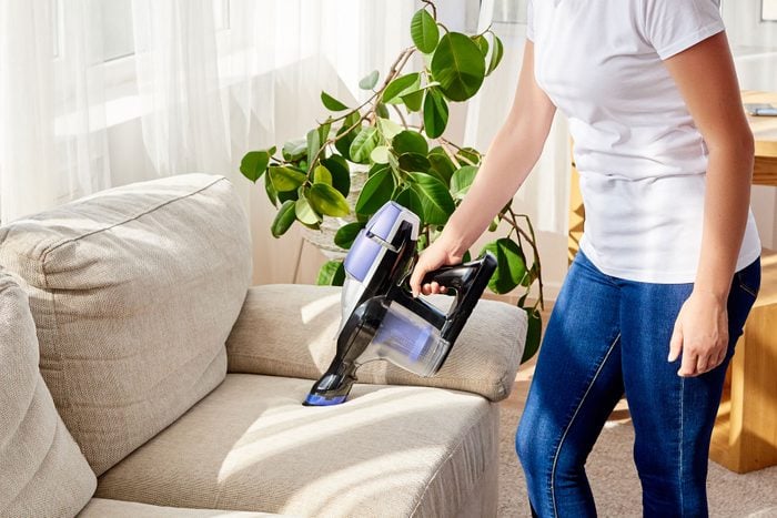 Close up of young woman in white shirt and jeans cleaning sofa with vacuum cleaner at home