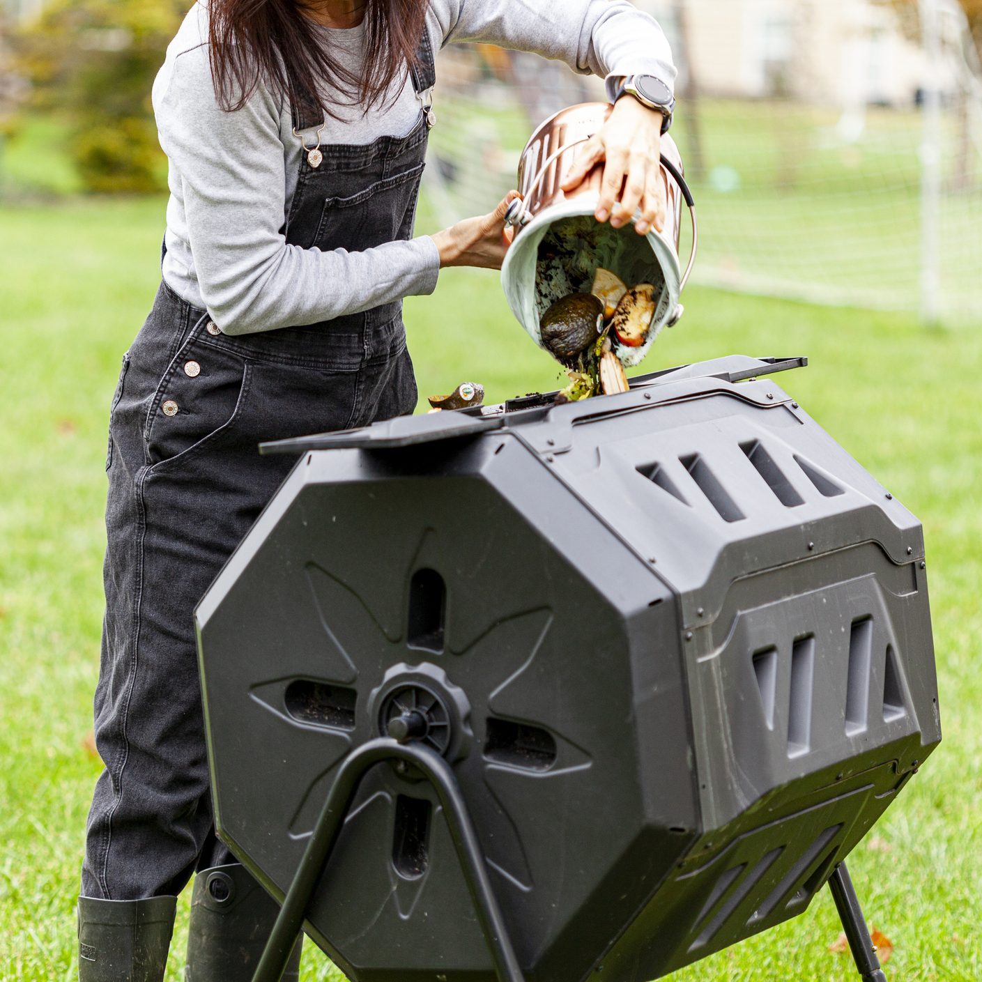 A woman is dumping a small bin of kitchen scraps into an outdoor tumbling composter in backyard garden