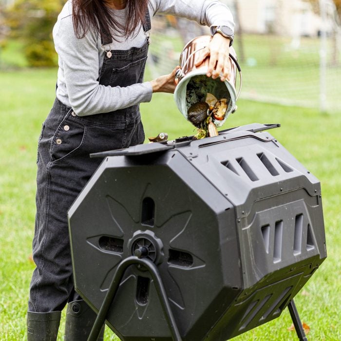 A woman is dumping a small bin of kitchen scraps into an outdoor tumbling composter in backyard garden